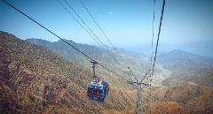 A cable car carrying passengers ascends through a mountainous landscape under a clear blue sky.