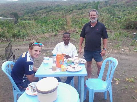 Three men are seated and standing around a blue table laden with food and drinks in an outdoor setting with a grassy background.