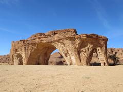 A massive, natural rock formation with archways resembling an elephant stands prominently in a sandy, desert landscape under a clear blue sky.