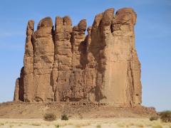 A large, natural rock formation with a reddish-brown hue rises prominently from a sandy desert landscape under a clear blue sky.