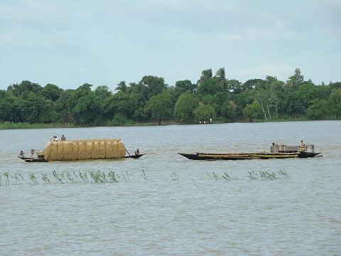 A wide shot of two boats carrying cargo and people across a large body of water, with lush green trees lining the distant shore.