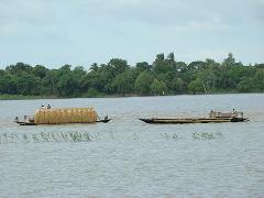 A wide shot of two boats carrying cargo and people across a large body of water, with lush green trees lining the distant shore.