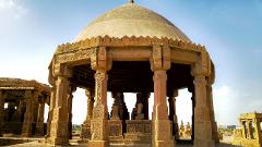 A stone domed structure with carved pillars stands in a field of similar monuments against a bright sky.