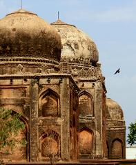 A worn, old mausoleum with multiple domes stands against a pale sky, showing its age and architectural details with a bird flying by.
