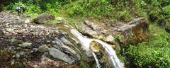 A scenic view of a small waterfall cascading down rocks surrounded by lush greenery, with two hikers walking along a rocky path in the background.