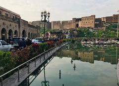 A scenic view of Erbil Citadel, a historic landmark, reflected in a pool with a pedestrian bridge in the foreground.