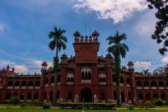 A full shot of Curzon Hall, a historical building in Dhaka, Bangladesh, with a red brick exterior, arched windows, and palm trees on either side, set against a blue sky with scattered clouds.