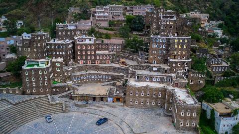 This is an aerial view of the historic village of Rijal Almaa in Saudi Arabia, showcasing its unique stone architecture and stepped landscape with modern cars parked below.