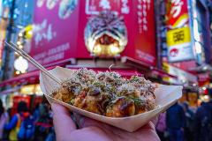A hand holds a boat-shaped tray of takoyaki, topped with bonito flakes and seaweed, in front of the vibrant Dotonbori street scene in Osaka, Japan.