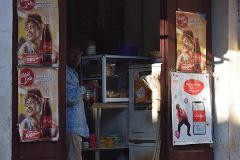 Free Daily Photography Walking Tour In Antananarivo's Historical City Center With Local Guide A man stands in the doorway of a shop, holding a small cup, with posters of World Cola and Airtel money displayed on the left and right sides.