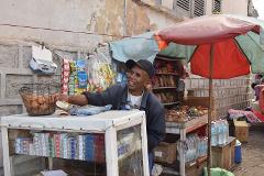 Free Daily Photography Walking Tour In Antananarivo's Historical City Center With Local Guide A smiling vendor stands at his stall in Antananarivo, Madagascar, with an assortment of goods including cigarettes, eggs, and bottled water.