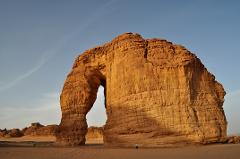 A majestic rock formation with a natural arch stands tall in a desert landscape under a clear sky, with a small figure of a person near its base emphasizing its immense scale.