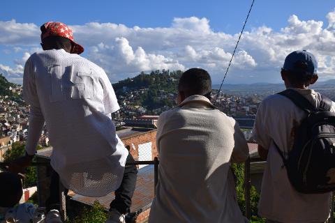 Free Daily Photography Walking Tour In Antananarivo's Historical City Center With Local Guide Three people stand on a balcony overlooking a densely populated city on a hill with a bright blue sky and clouds.