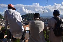 Free Daily Photography Walking Tour In Antananarivo's Historical City Center With Local Guide Three people stand on a balcony overlooking a densely populated city on a hill with a bright blue sky and clouds.