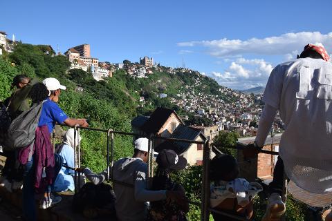 Free Daily Photography Walking Tour In Antananarivo's Historical City Center With Local Guide A group of people stand on a balcony overlooking a densely populated hillside city with buildings of various sizes and colors, under a bright blue sky with scattered clouds.
