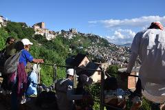 Free Daily Photography Walking Tour In Antananarivo's Historical City Center With Local Guide A group of people stand on a balcony overlooking a densely populated hillside city with buildings of various sizes and colors, under a bright blue sky with scattered clouds.