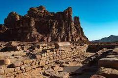 Okay, here's a description of the image in one sentence and under 320 characters:The image shows ancient ruins of a stone structure with a rocky mountain backdrop against a clear blue sky, likely located in a desert region.