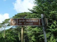 A sign reading "Jardin Botanique de Coconi" stands amidst lush green trees under a partly cloudy sky.