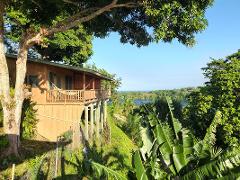 A stilt house with a deck and hammock overlooks a lush, tropical landscape with a glimpse of blue water in the distance under a clear blue sky.