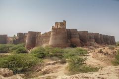An ancient fort with cylindrical towers and high walls stretches across the landscape, surrounded by lush greenery and sandy terrain, under a hazy sky.