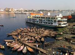 A bustling river scene with numerous small boats clustered near a large ferry named "M.V. PRINCE AWLAD-4" against a cityscape backdrop.