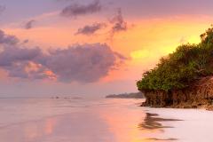 A vibrant sunset paints the sky with hues of orange, pink, and purple over a serene beach scene, featuring tranquil waters, a sandy shore, and a rocky, tree-covered headland.