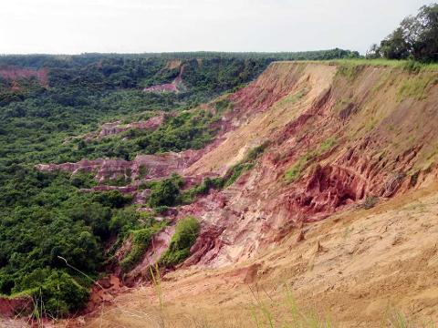 A dramatic landscape of a steep, eroded cliff face with reddish-pink rock layers and lush green vegetation at its base.