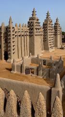 This is a vertical, eye-level shot of the Great Mosque of Djenné in Mali, showcasing the towering mud-brick structure with its distinctive wooden scaffolding and pointed minarets under a clear sky.