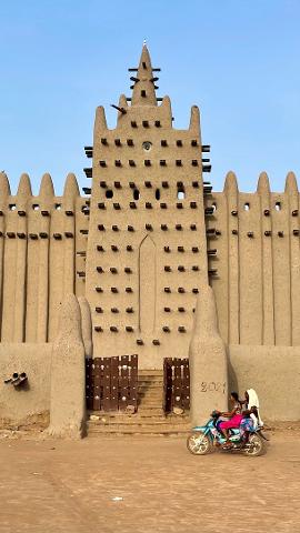 A stunning view of the Great Mosque of Djenné, with its mud-brick architecture and distinctive wooden scaffolding, as two people ride by on a scooter.