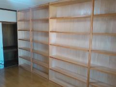 A wide wooden bookshelf with many shelves stands against a light-colored wall in a room with a wooden floor.