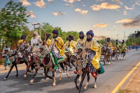 A group of people in traditional attire are riding horses down a road, possibly as part of a cultural event or parade.