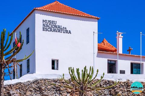 The Museu Nacional da Escravatura, a white building with a red tiled roof, stands against a bright blue sky, with cacti in the foreground and a stone wall at its base.