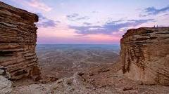 Here's a description of the image in one sentence:A wide, panoramic view of a desert landscape framed by two rocky cliffs under a colorful sunset sky, showcasing a vast, arid expanse.