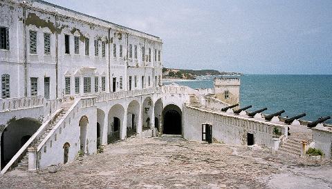 This is a view of Cape Coast Castle, a white-walled building complex with cannons along its perimeter, located on the coast of Ghana.