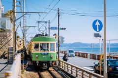 A green and yellow train, marked with "305", travels along tracks near the coast, with a person and vehicles nearby, and signage indicating "Kamakura koko Sta.".
