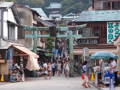 A bustling street scene in Japan features a large torii gate framing a crowd of people walking up a hill, lined with shops and restaurants under various awnings and umbrellas, with a temple visible in the distance.