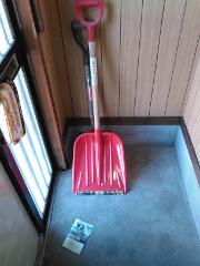 A red snow shovel stands upright against a wooden wall, next to a window with blinds and a brochure on the floor.