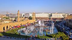 A vibrant city scene shows a bustling square with fountains and people, set against a backdrop of buildings and clear blue sky.