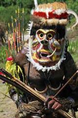A person in traditional attire, likely for the Rabaul Mask Festival, wears a colorful, intricate mask and carries a quiver of arrows.