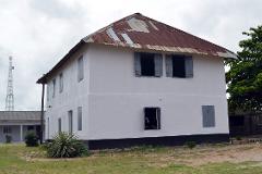A two-story white building with a rusty corrugated metal roof sits on a grassy lawn under a cloudy sky.