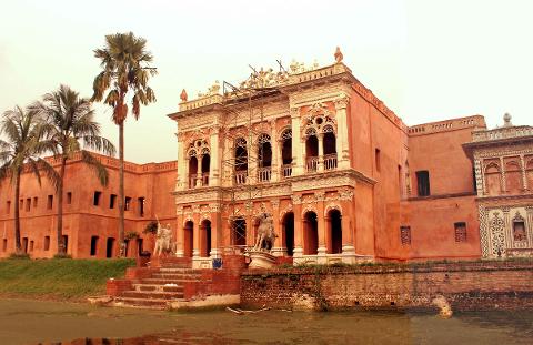 5-Day Dhaka Cultural Heritage Tour: Floating Markets & River Life Experience A grand, terracotta-colored palace with ornate architecture stands beside a tranquil, murky body of water, framed by lush green grass and tall palm trees.