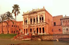 5-Day Dhaka Cultural Heritage Tour: Floating Markets & River Life Experience A grand, terracotta-colored palace with ornate architecture stands beside a tranquil, murky body of water, framed by lush green grass and tall palm trees.