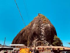 Gran Tour incluyendo Bogotá, Medellín, Nuquí y Valle del Cocora - 11 Días A large rock formation with a winding staircase leading to the top is surrounded by vendors, trees, and a clear blue sky.