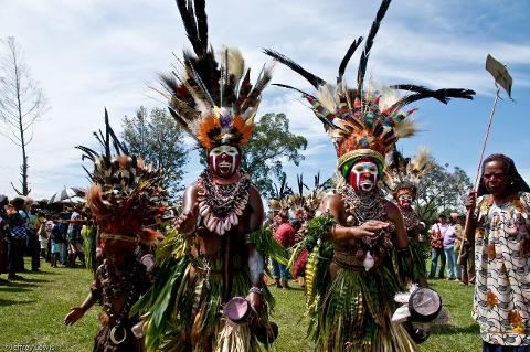 A vibrant scene from the Goroka Show Festival in Papua New Guinea showcases performers adorned with elaborate feather headdresses, facial paint, shell necklaces, and grass skirts.
