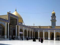 A large mosque with a golden dome and a clock tower sits in a courtyard with people walking around.