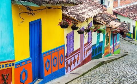A vibrant streetscape showcases colorful houses with tiled roofs, adorned with hanging flower baskets and intricate painted details, all set against a cobblestone street.