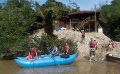 A group of people are preparing for a rafting adventure on a river near a rustic wooden building with stairs.