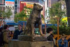 A bronze statue of a Shiba Inu dog stands prominently in a bustling Tokyo intersection, surrounded by people and illuminated by bright city lights and signage.
