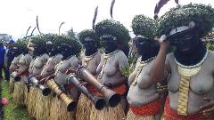 A line of women, painted with black faces, wearing leaf headdresses and grass skirts, hold long wooden instruments in an outdoor setting.