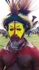 Mt Hagen & Paiya Show Package A man wearing a colorful headdress and tribal paint, with a yellow face and red accents, stands out in a crowd at the Mount Hagen Festival & Paiya Show.